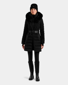 A person wearing the Rudsak ALISA M - L125586 Black fitted quilted down parka with fur-trimmed hood, black pants, knee-high boots, and a black beanie stands against a plain light background.
