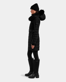 A woman wears the Rudsak ALISA M - L125586 Black parka with a belted waist and fur-trimmed hood, styled with a black beanie, leggings, and knee-high boots, standing in profile against a plain light background.