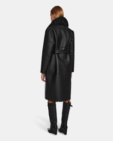 A woman with straight, light brown hair is seen from behind wearing the Rudsak ANNABELLE - L124544 Black vegan leather coat with a shearling collar, belted waist, and knee-high black boots against a plain white background.