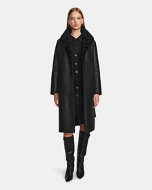A woman faces forward, wearing the Rudsak ANNABELLE - L124544 Black vegan leather coat over a black dress and knee-high boots, set against a plain white background.