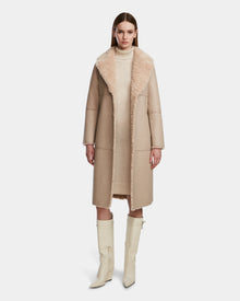 A woman faces forward, wearing the Rudsak ANNABELLE - L124544 Sand reversible teddy coat with a large fuzzy collar over a light knit dress and knee-high off-white boots. The background is plain white.