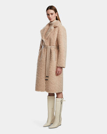 A woman wearing the Rudsak ANNABELLE - L124544 Sand belted faux shearling coat and cream knee-high heeled boots stands against a plain white background, looking forward with a neutral expression.
