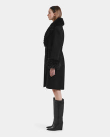 A woman stands in profile wearing the ARTIS - L125575 Black women’s wool coat by Rudsak, featuring a faux fur collar and cuffs, belted waist, black knee-high boots, and a black headband against a plain white background.