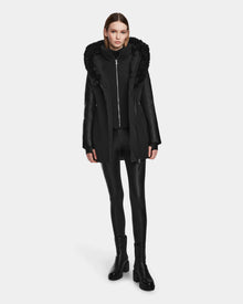A woman stands facing forward against a white background, wearing the Rudsak AURELLE - L124538 Black hooded jacket with faux fur trim and vegan leather sleeves, styled with black leggings and heeled boots for winter protection.