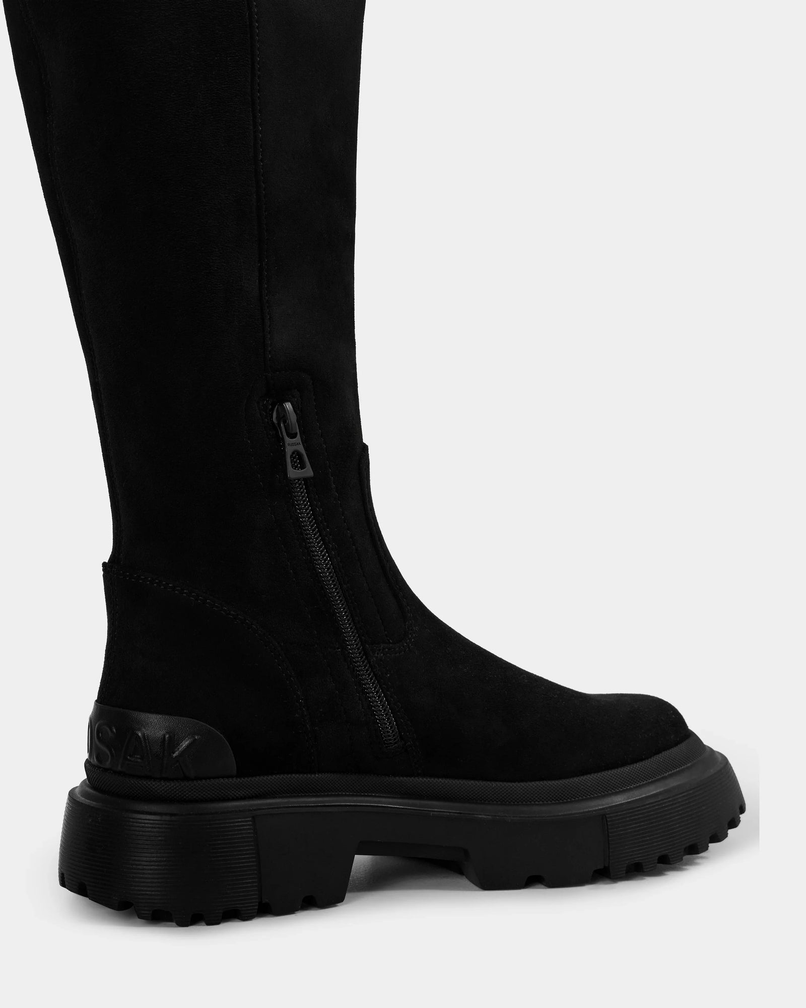 A close-up side view of the AVOLA - L225514 Black women’s knee-high boot by Rudsak in faux suede, featuring a chunky lug sole, side zipper, and embossed branding on the heel, shot against a plain light background.