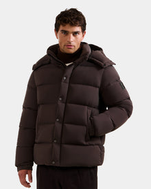 A man stands with one hand in his pocket against a plain background, wearing the Rudsak BRAYDEN M-S - M125524 Espresso down puffer jacket, featuring a hood and snap buttons.