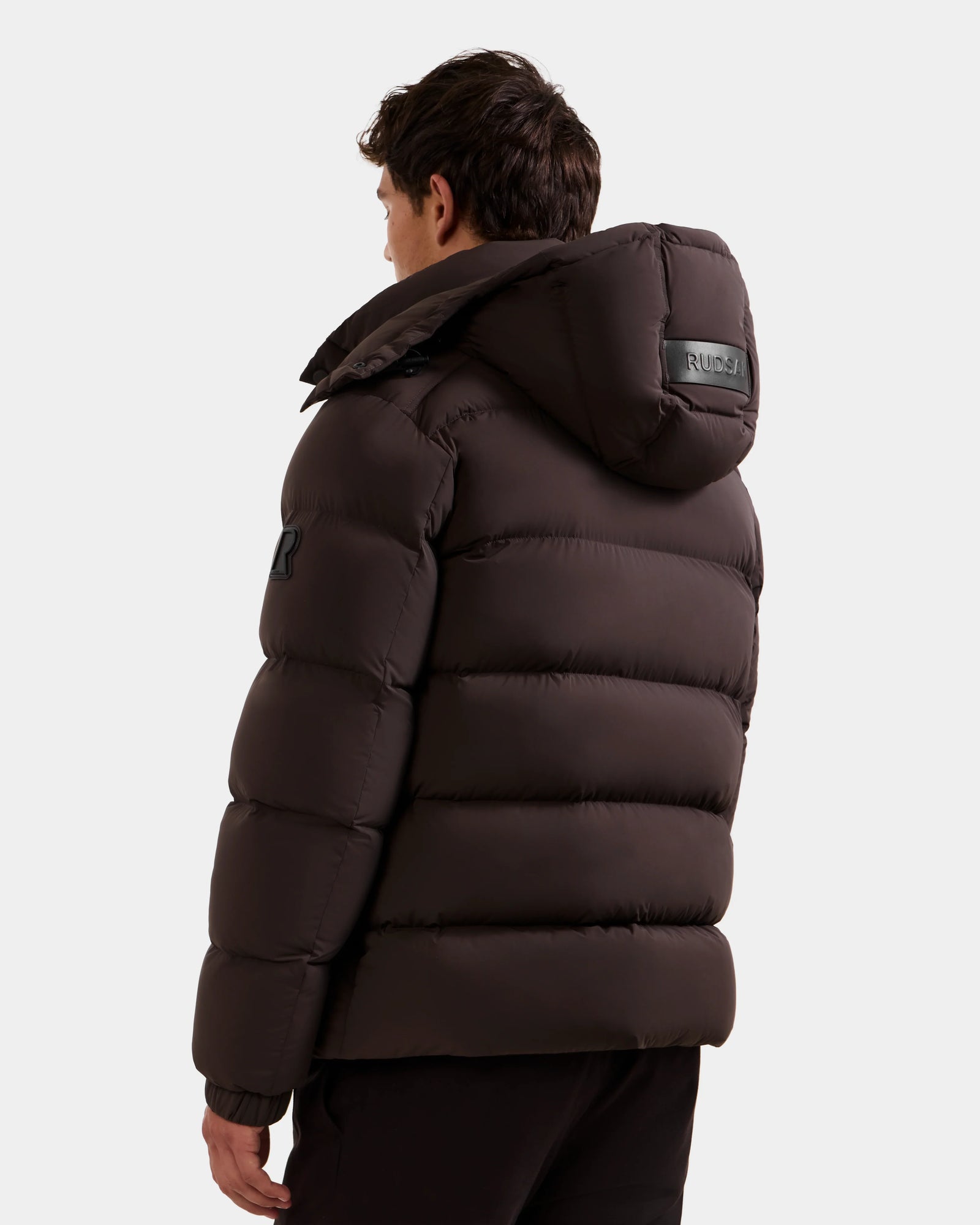 A person with dark hair is shown from the back, wearing the Rudsak BRAYDEN M-S - M125524 Espresso men’s down puffer jacket featuring a rubber patch on the upper sleeve and a label on the hood, against a plain light background.