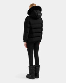 A person stands with their back turned, wearing the Rudsak BREE M - L125582 Black women’s down puffer jacket with a fur-trimmed removable hood, black leggings, and winter boots—an essential cold-weather look on a white background.