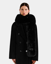 Wearing the Rudsak CARMY - L425534 Black long double-breasted coat and a plush women’s faux-fur scarf, a person stands against a plain light background, looking directly at the camera with a neutral expression.