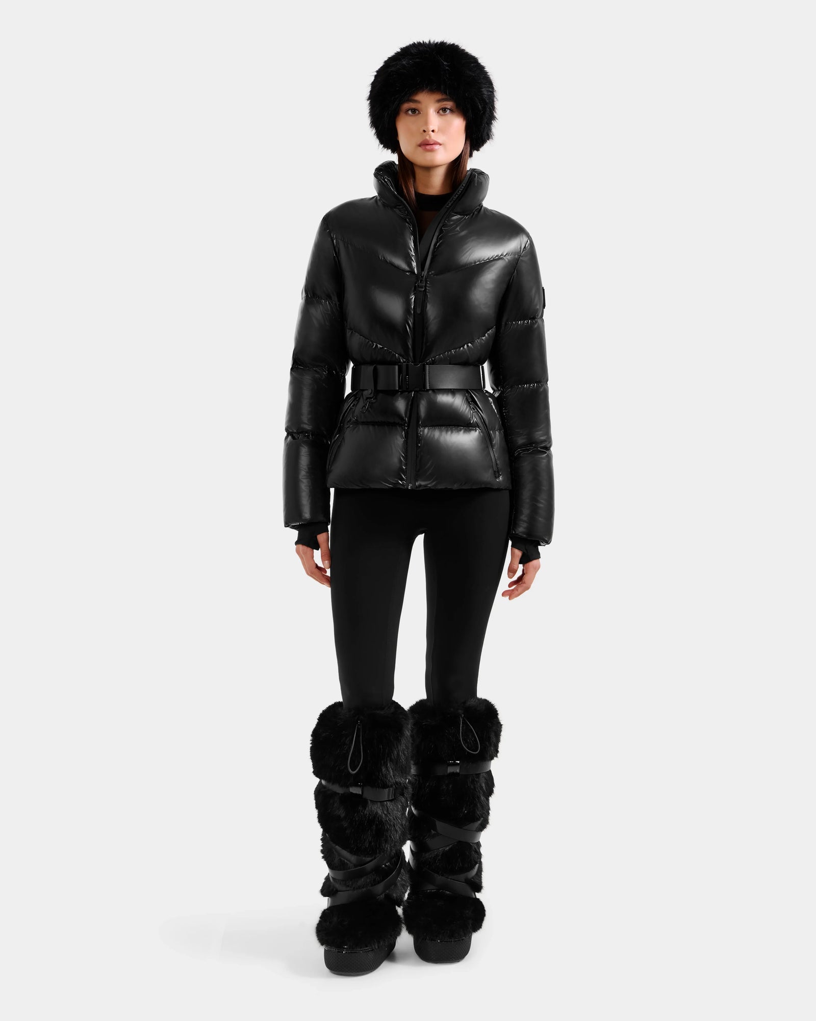 A woman faces forward against a white background, wearing the Rudsak CHRISTELLE S - L125544 Black belted women’s down puffer jacket with faux fur trim, paired with black leggings, a black fur hat, and black furry boots.