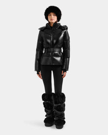 A person stands facing forward, wearing the Rudsak CHRISTELLE S - L125544 Black women’s down puffer jacket with a belt, paired with black leggings, furry boots, and a faux fur trim hat against a plain light background.