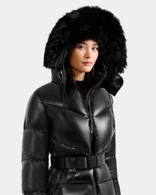 A woman wears the Rudsak CHRISTELLE S - L125544 Black down puffer with a large faux fur trim hood and black belt, standing against a plain light background.