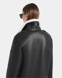 Seen from behind against a plain light background, a person with long, straight brown hair wears black sunglasses and the Rudsak DARCY - L125539 Black vegan leather overcoat with a white shearling collar.