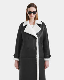 A person models the Rudsak DARCY - L125539 Black women's faux sherpa coat with white trim, standing against a plain white background and looking straight ahead with a neutral expression.