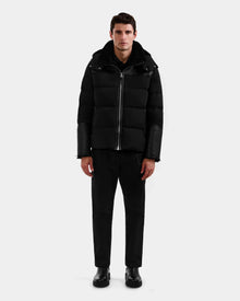 A man stands facing forward against a plain light gray background, wearing the Rudsak DAVID - M125508 Black men’s shearling down puffer with leather sleeves, paired with black pants and black shoes.