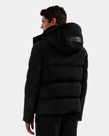 A man with short dark hair wears the Rudsak DAVID - M125508 Black puffer jacket, featuring a visible RUDSAK logo on the back. This men's shearling down puffer is winter-ready, and he faces away from the camera against a plain background.