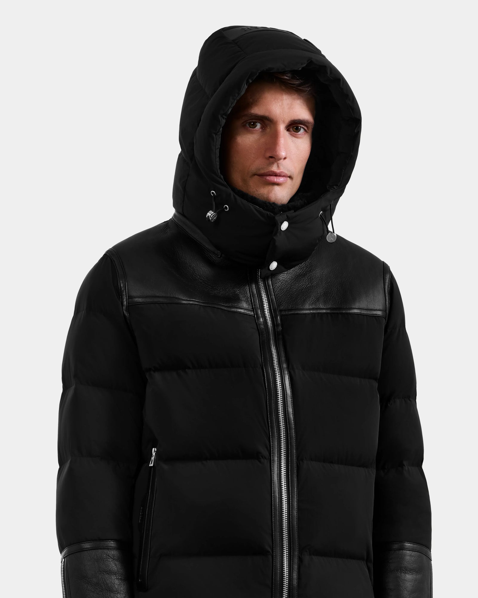 A man stands facing forward against a plain light background, wearing the Rudsak DAVID - M125508 Black men’s shearling down puffer with leather accents, a hood, front zipper, and button details.