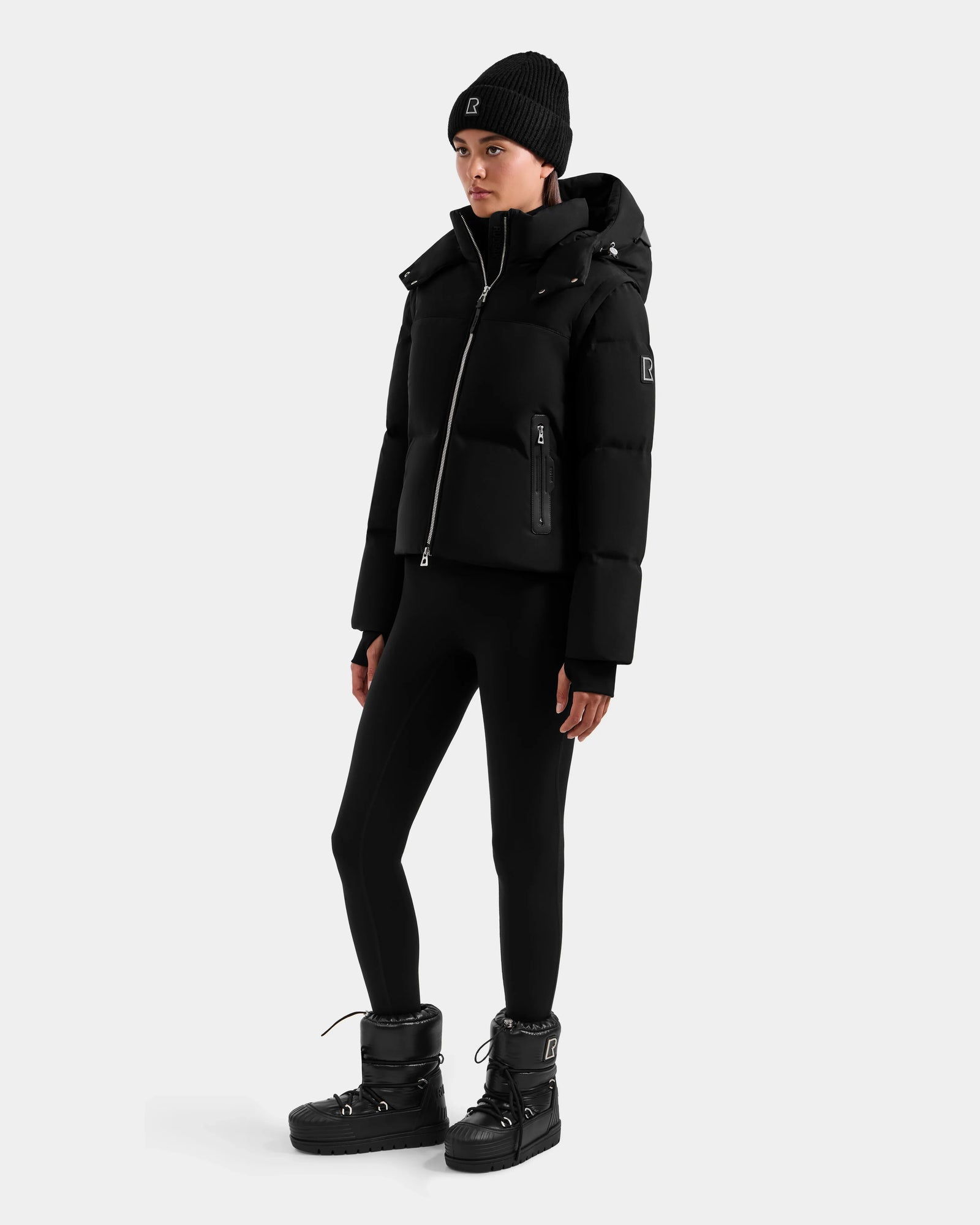 A woman wears the Rudsak DELILAH M - L125538 Black down puffer with removable sleeves, black leggings, winter boots, and a black beanie, standing against a plain light gray background.