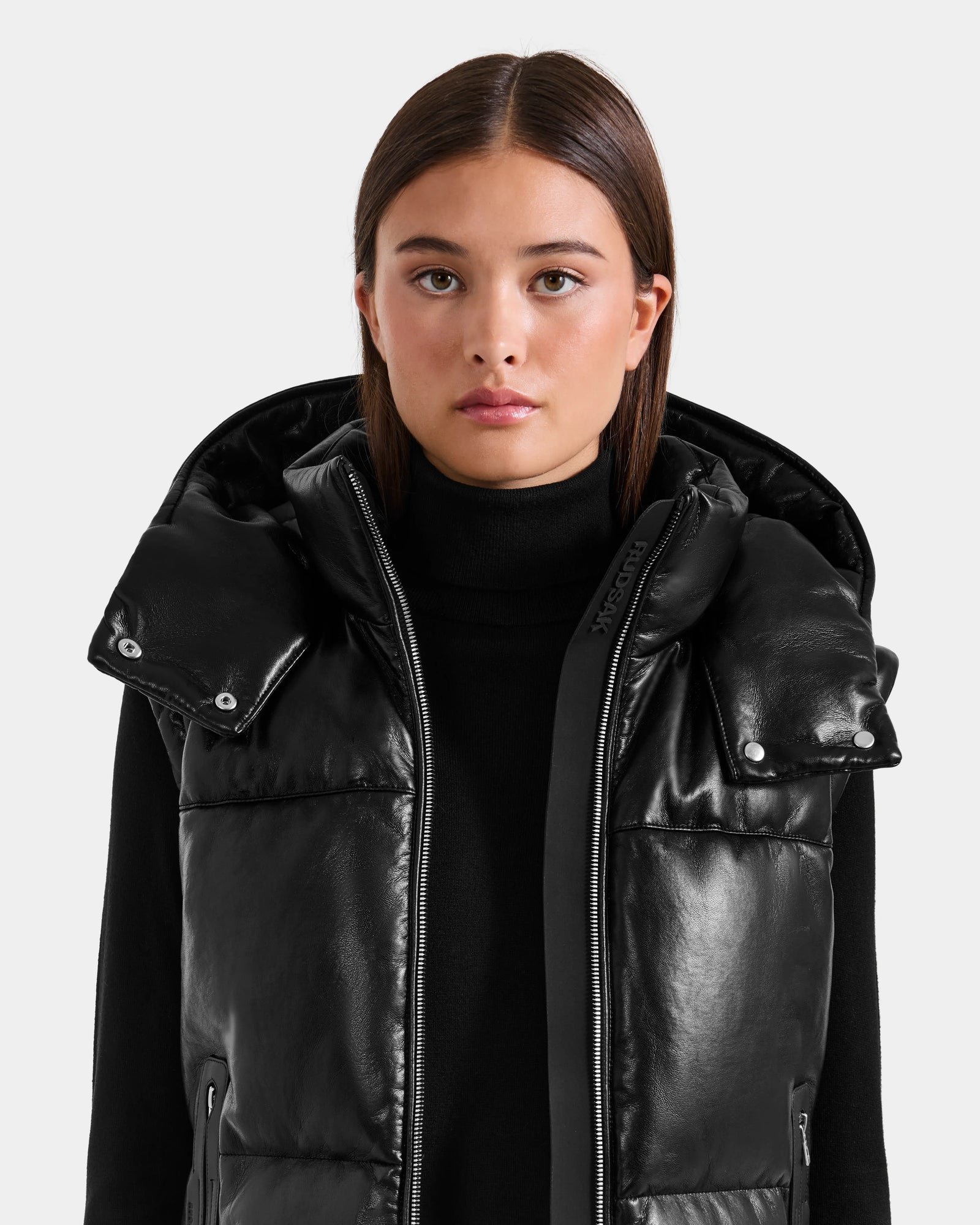 A woman with straight brown hair models the Rudsak DELILAH PX - L125537 Black vegan-leather puffer vest with a large collar and a black turtleneck, standing against a plain light gray background.