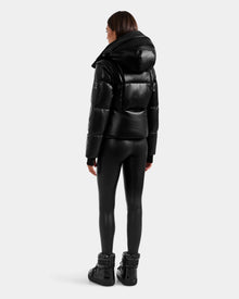 A person stands facing away, wearing the Rudsak DELILAH PX - L125537 Black vegan-leather puffer jacket, black fitted leggings, and black high-top shoes against a plain light gray background.