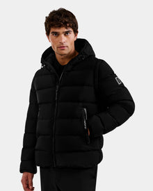 A man with short brown hair wears the Rudsak DEMIAN M-R - M123504 Black men's down puffer jacket, featuring horizontal quilting and crafted from sustainable recycled fabric. He stands facing forward with one hand in his pocket against a light gray backdrop.