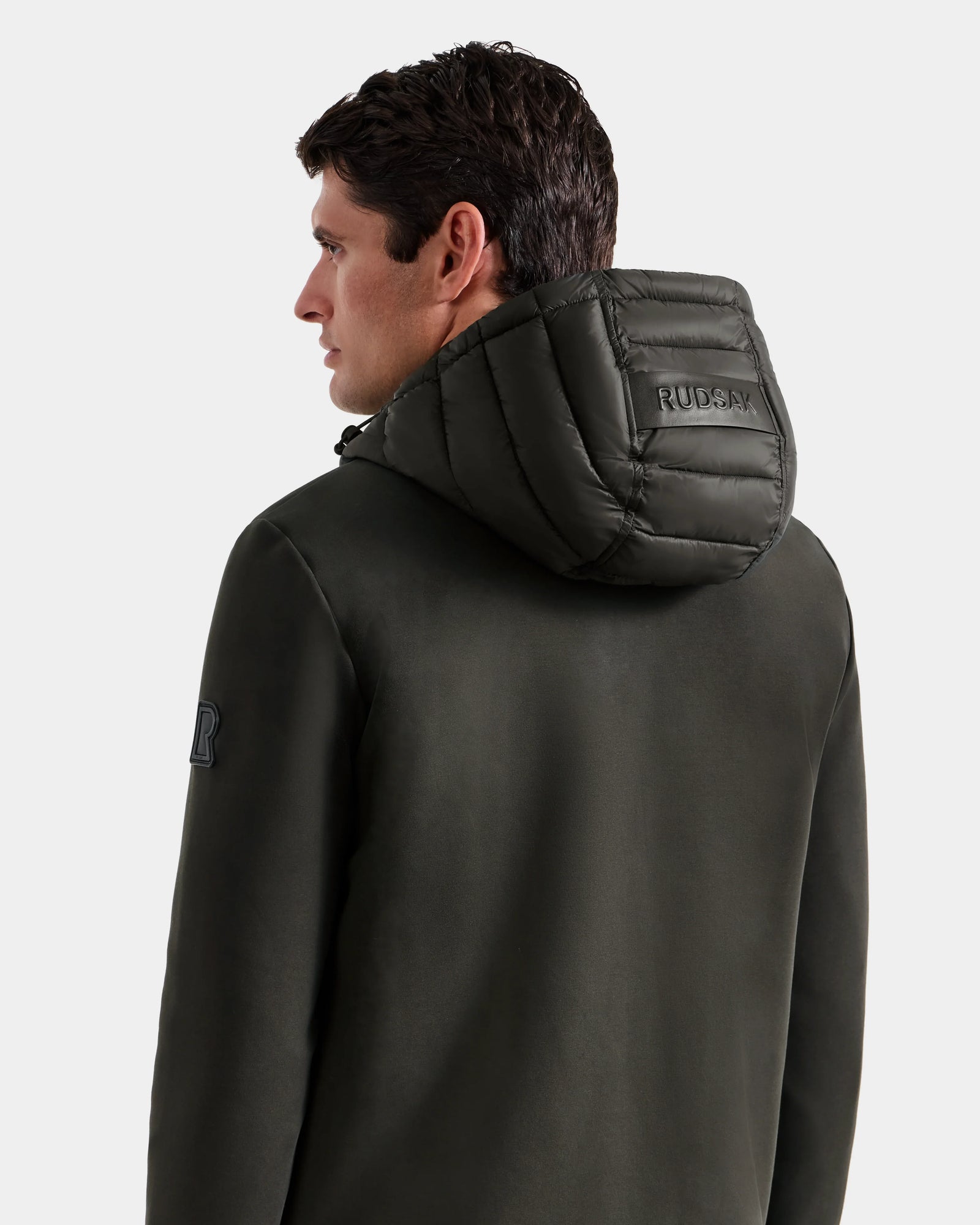 A man with short brown hair is seen from behind wearing the Rudsak DURIN M - M125530 dark olive men’s hooded jacket, which features visible stitching, Rudsak branding on the hood, a logo patch on the left sleeve, and waterproof zippers.
