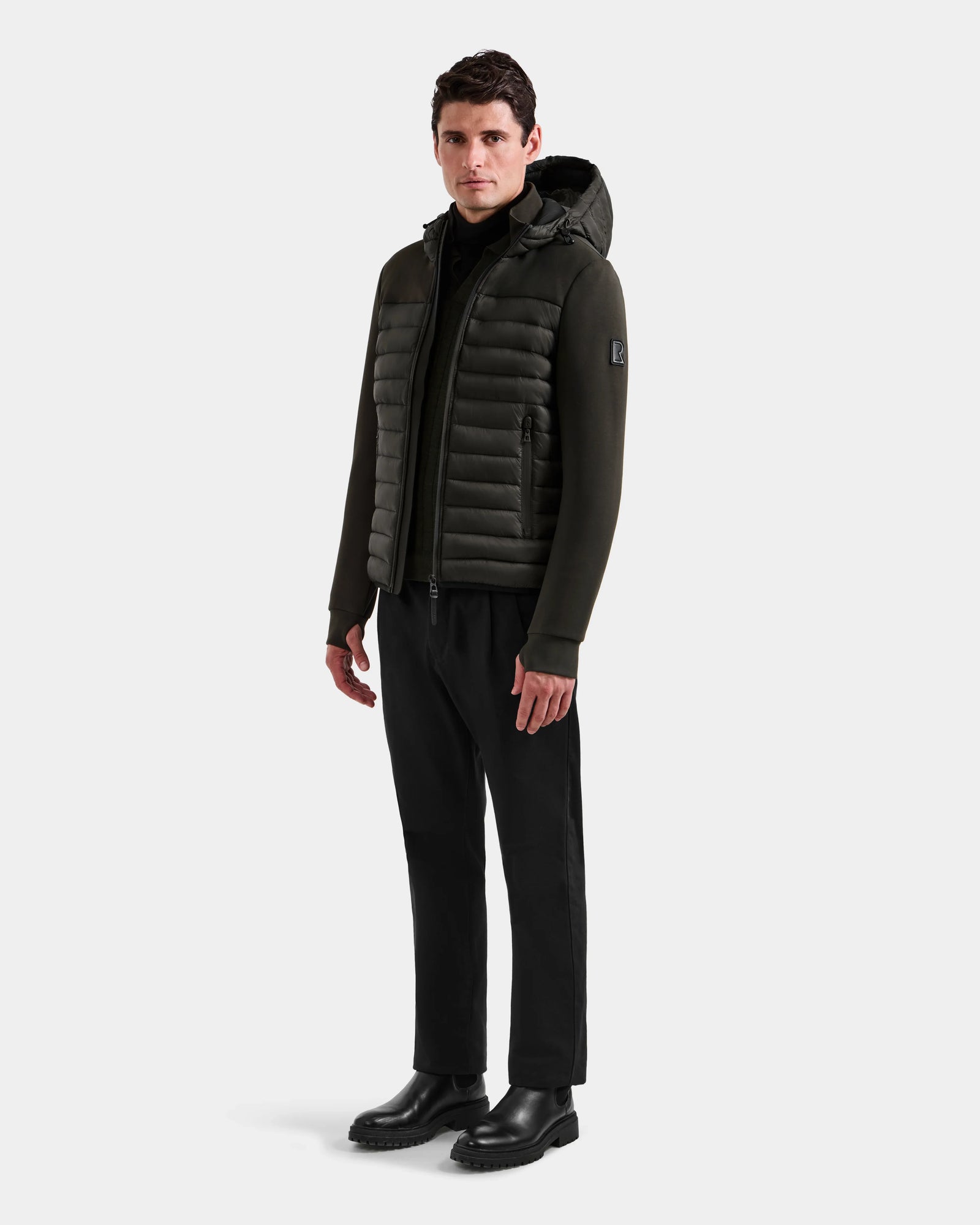 A man stands facing forward, wearing the Rudsak DURIN M - M125530 Dark Olive men's hooded jacket, paired with black pants, a black turtleneck, and black boots against a plain light background.