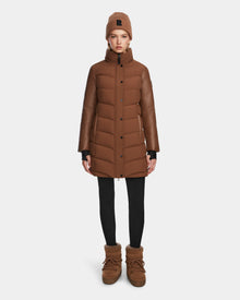 A woman faces forward against a white background, wearing the Rudsak ELSA - L123535 Taupe long quilted down puffer coat, a matching brown knit beanie, black leggings, and brown winter boots.
