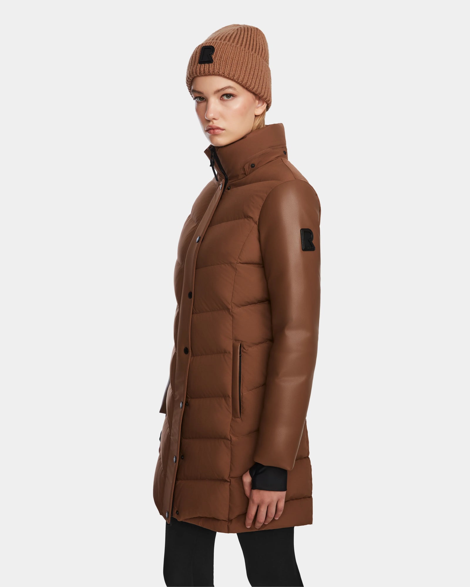 A woman in the Rudsak ELSA - L123535 Taupe long quilted down puffer with faux leather sleeves and a brown knit beanie stands sideways against a plain light background.