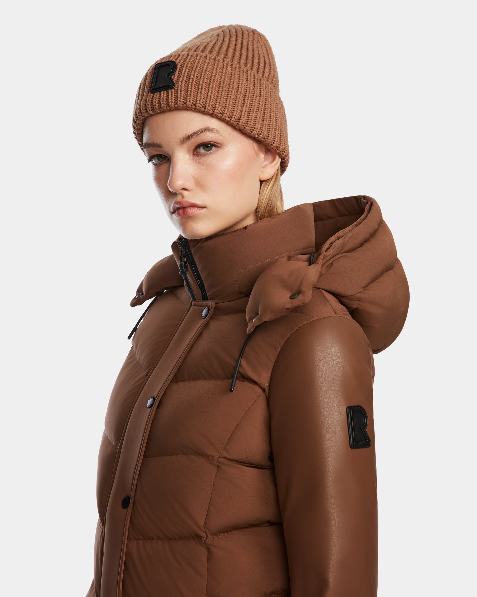 A woman wearing the Rudsak ELSA - L123535 Taupe quilted down puffer jacket with a hood and a brown knit beanie stands against a plain white background, looking slightly to the side with a neutral expression.