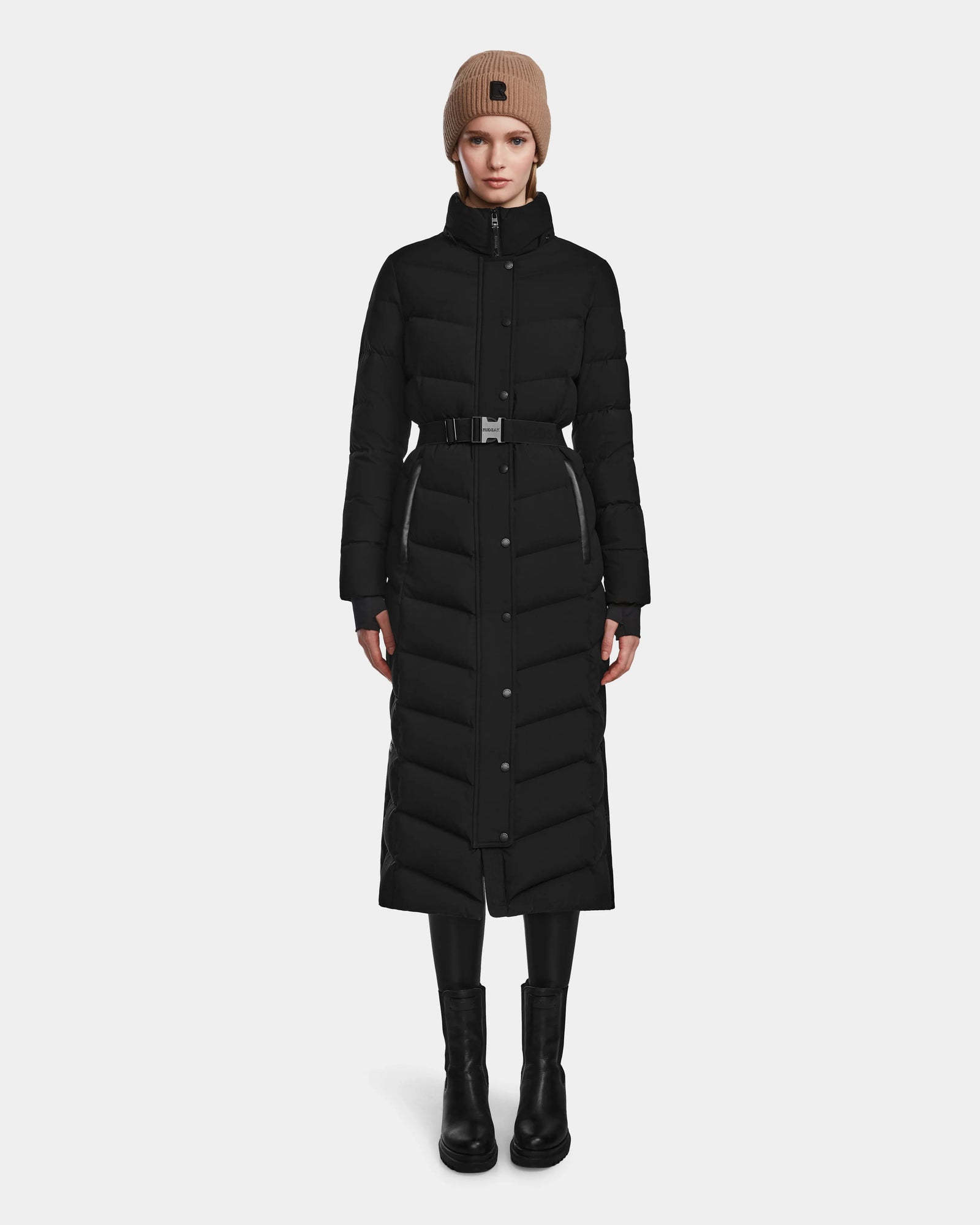 A person wears the Rudsak ELSIE M - L123517 Black/Natural women’s maxi down puffer with a belt, paired with black leggings, boots, and a tan knit beanie. The RDS-certified coat offers both warmth and sustainable style against a plain light background.