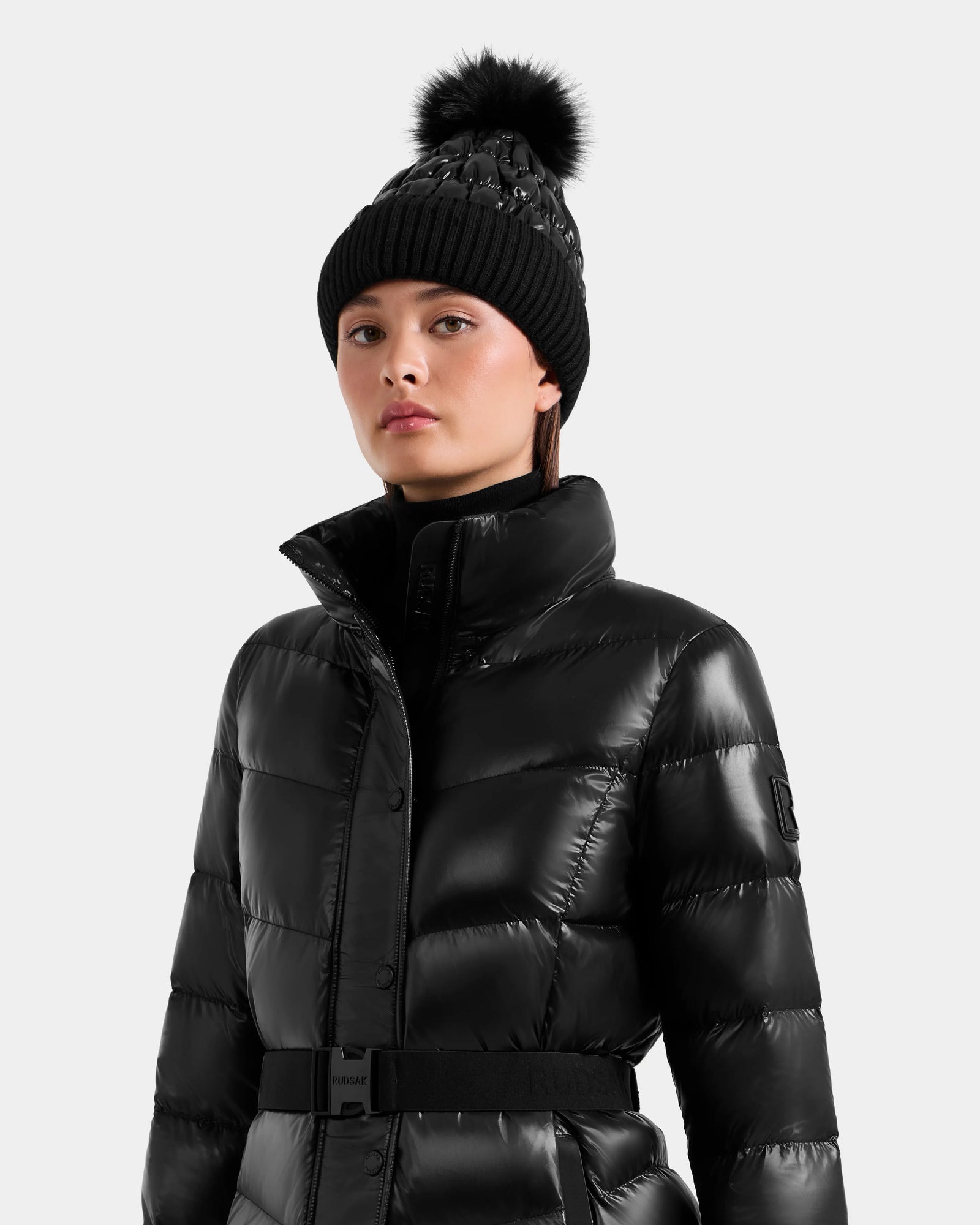 A woman wears the Rudsak ELSIE S - L124536 Black shiny water-resistant puffer with high collar and belted waist, paired with a black pom-pom knit beanie, standing against a plain light gray background.