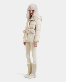 A woman stands against a white background wearing the Rudsak EMBER M-M - L125524 down puffer in Vanilla, featuring RDS-certified down, fur trim, a matching hat, slim pants, and tall beige boots.