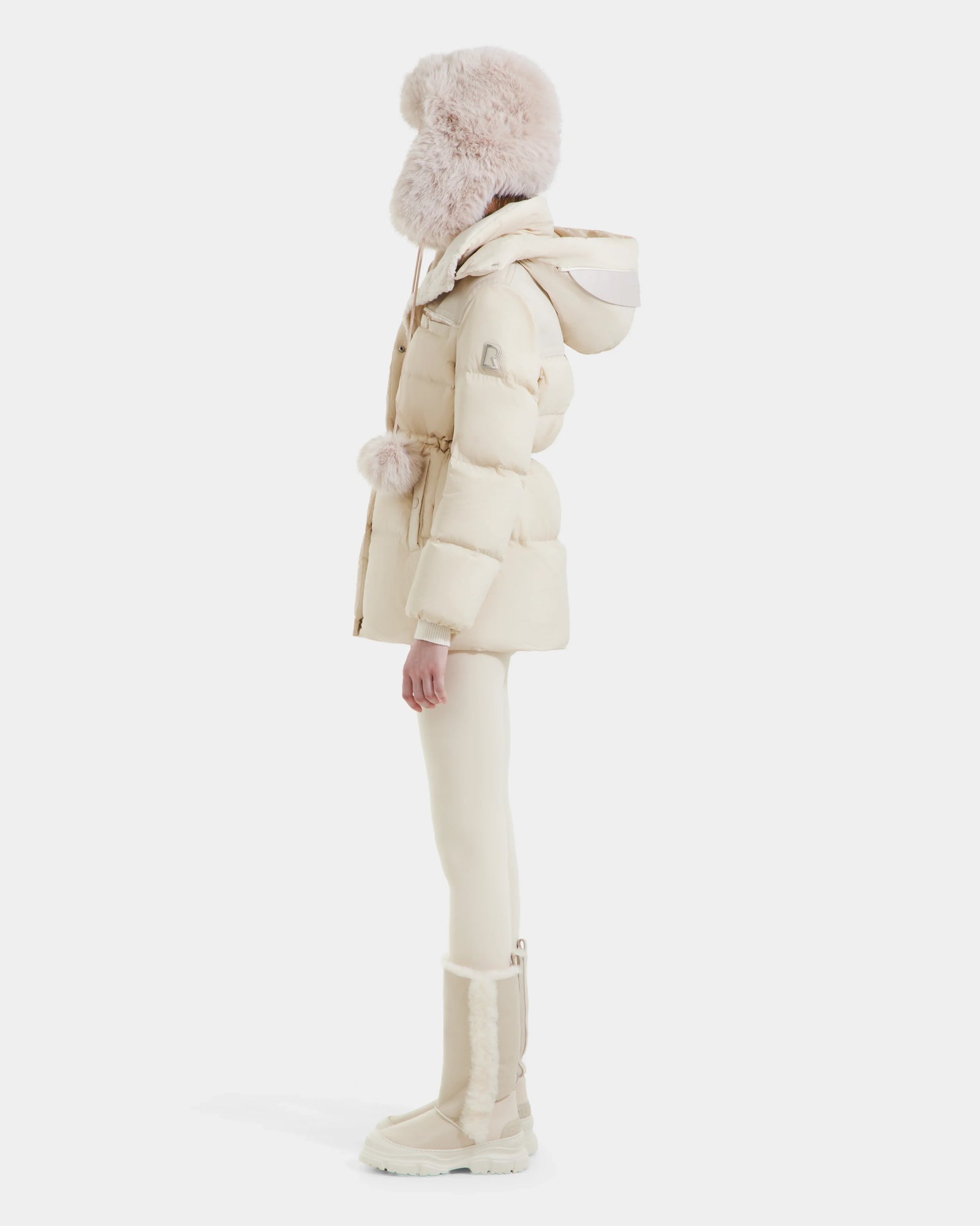 A person in profile wears the Rudsak EMBER M-M - L125524 Vanilla vegan-leather puffer jacket with a hood, paired with fitted pants, tall boots, and a large fluffy hat. Their all-cream winter outfit contrasts against a white background.
