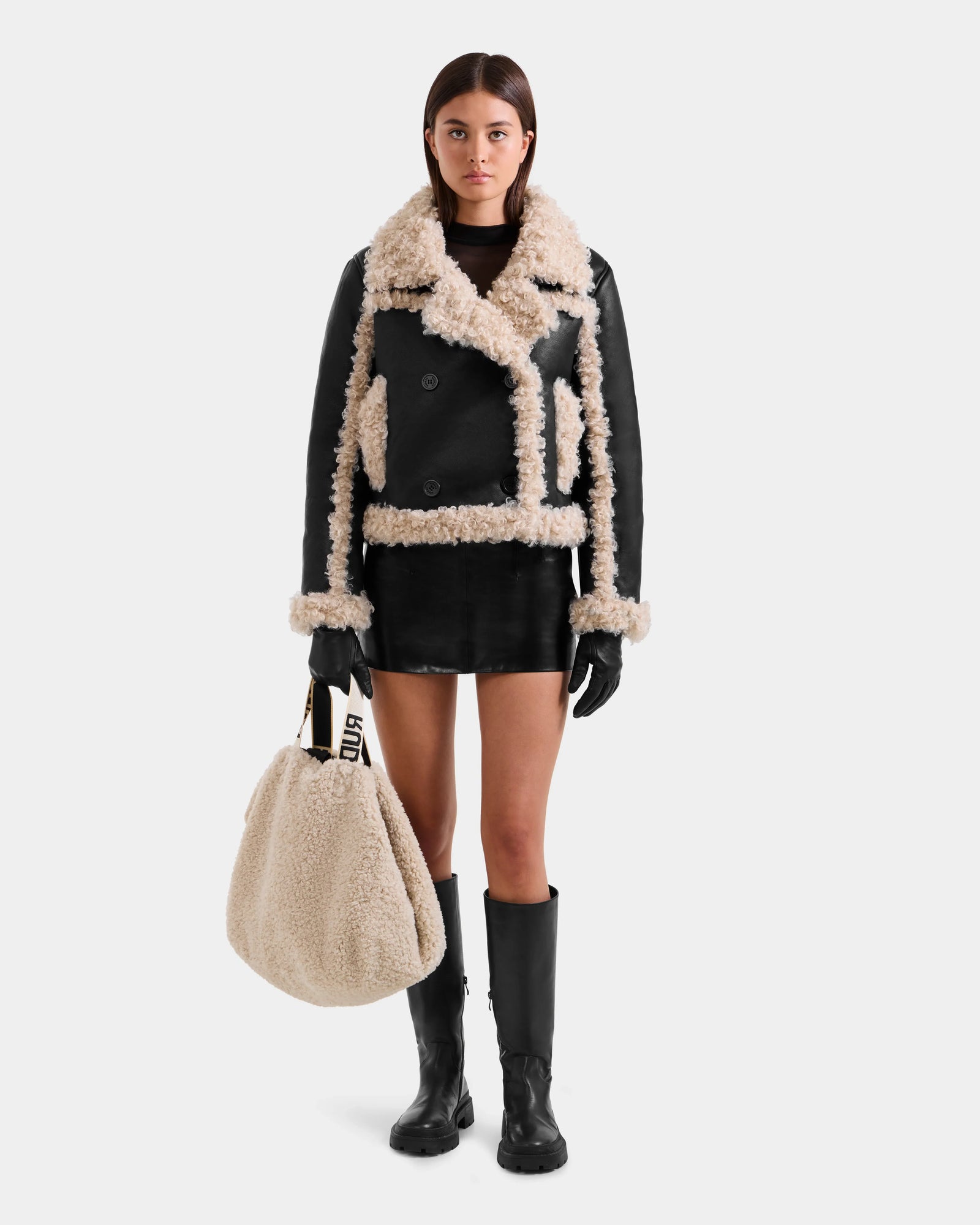 A woman stands facing forward in the Rudsak EMERY - L125548 Black vegan-leather jacket with beige faux shearling trim, paired with a black mini skirt, gloves, knee-high boots, and holding a large beige shearling bag against a white background.