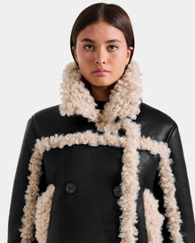A woman with straight brown hair wears the Rudsak EMERY - L125548 Black vegan-leather jacket with cream faux shearling trim and large buttons, looking directly at the camera against a plain light background.