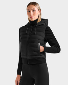 A woman models the Rudsak EMILY - L125515 Black hooded down bomber over a ribbed long-sleeve top and leggings, standing confidently against a plain background to highlight its cropped modern silhouette.