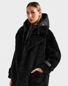 A woman wears the Rudsak EMRYN - L125588 Black textured teddy jacket with a hood, one hand in her pocket, against a plain light background.