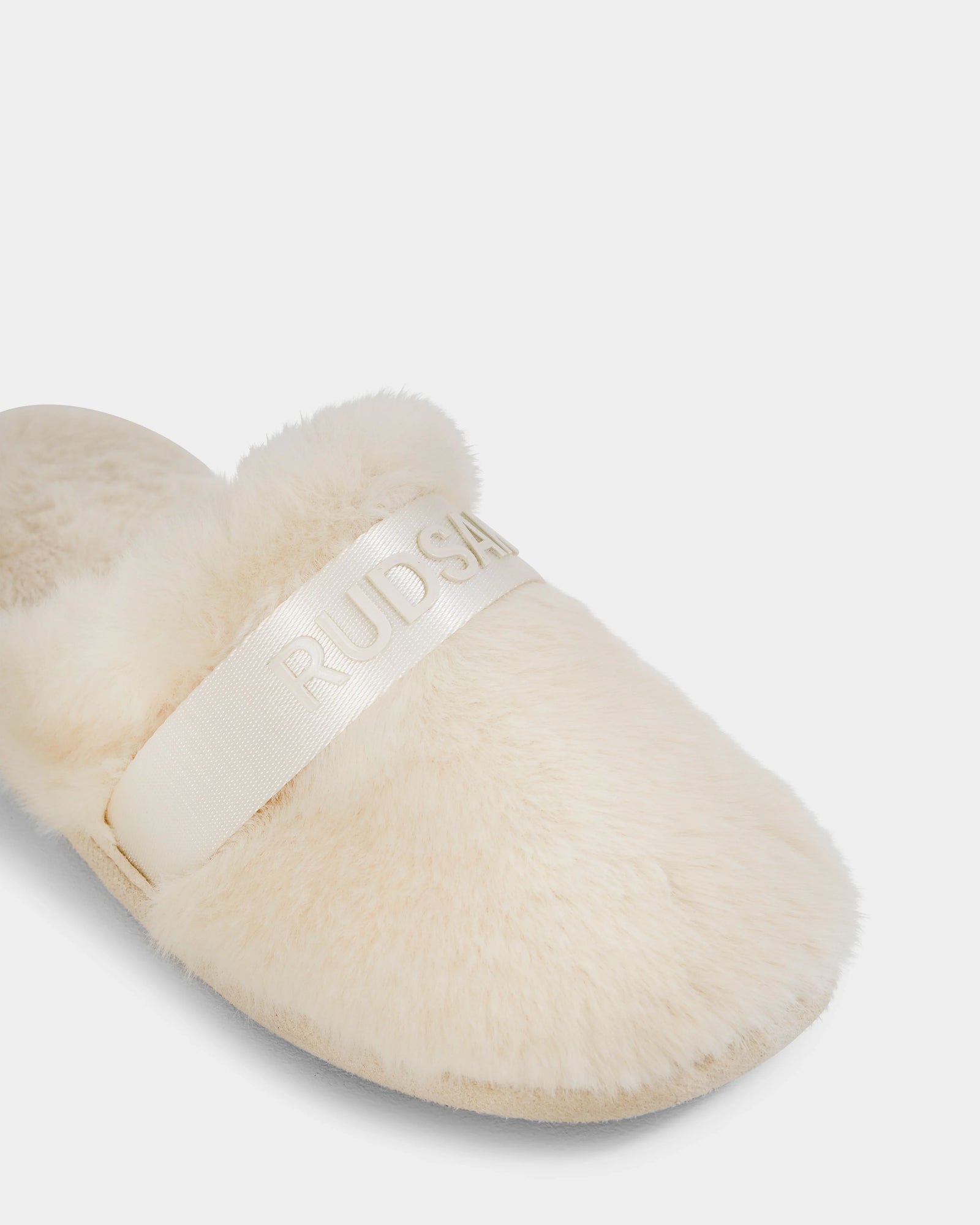 A close-up of the FLUFF - L223509 Cream women's slipper by Rudsak, featuring cream-colored faux fur and a wide shiny band with embossed RUDSAK text, set against a plain white background for a cozy lounge look.
