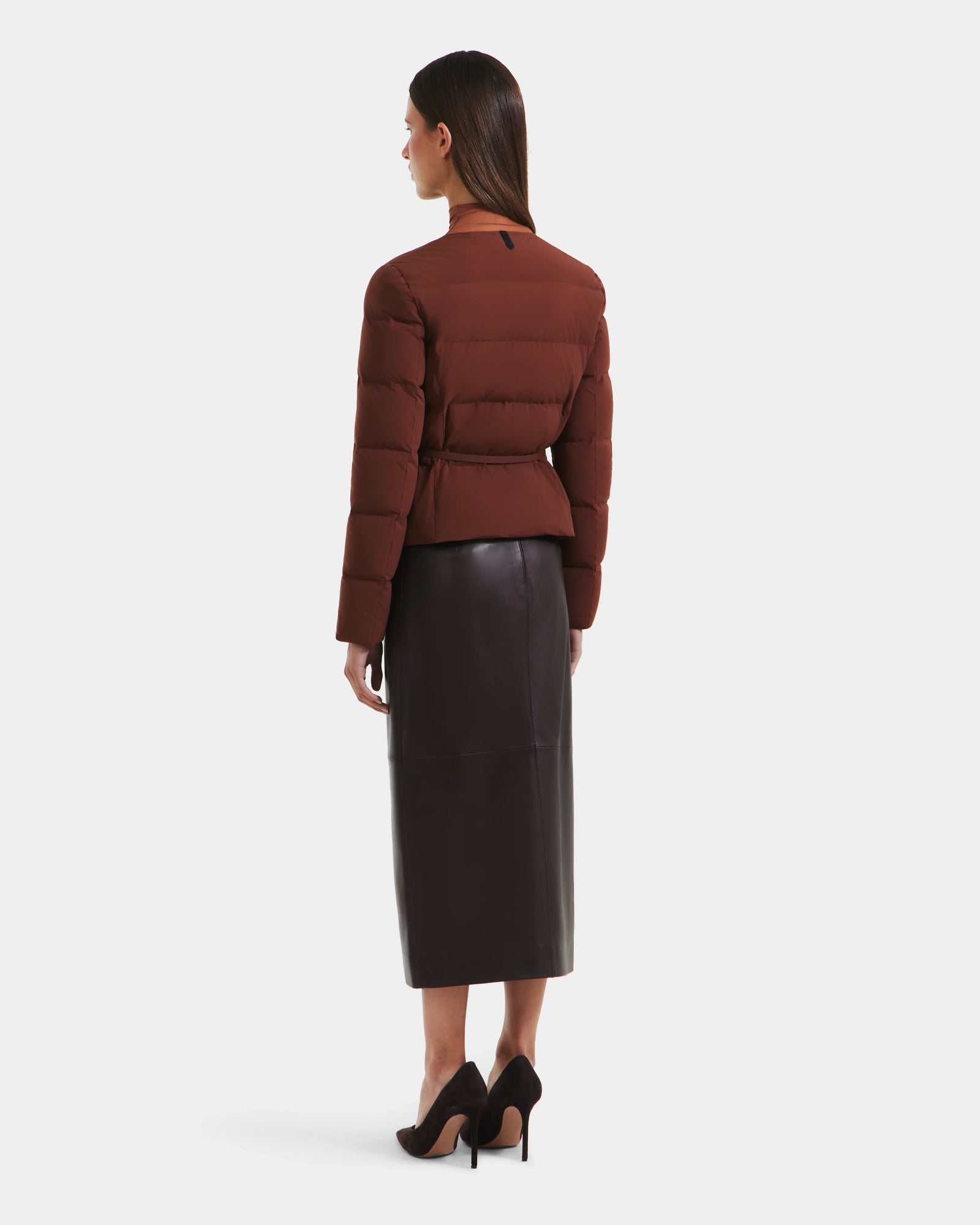 A woman faces away in the Rudsak FRANCESCA - 8125021 Rust puffer jacket, paired with a black midi leather skirt and black high heels, set against a plain white background.