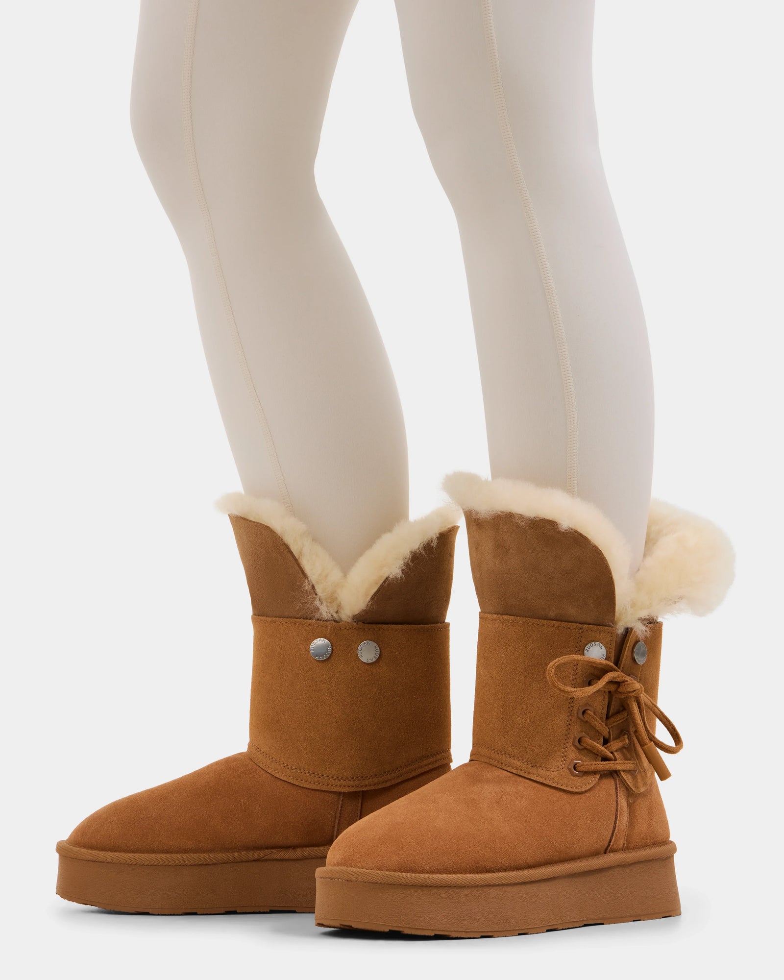 A person wears cream tights and cozy winter boots—one is the Rudsak INESA - L225026 Tan women’s suede platform boot with wool lining and decorative buttons, while the other has a lace-up design for added warmth.