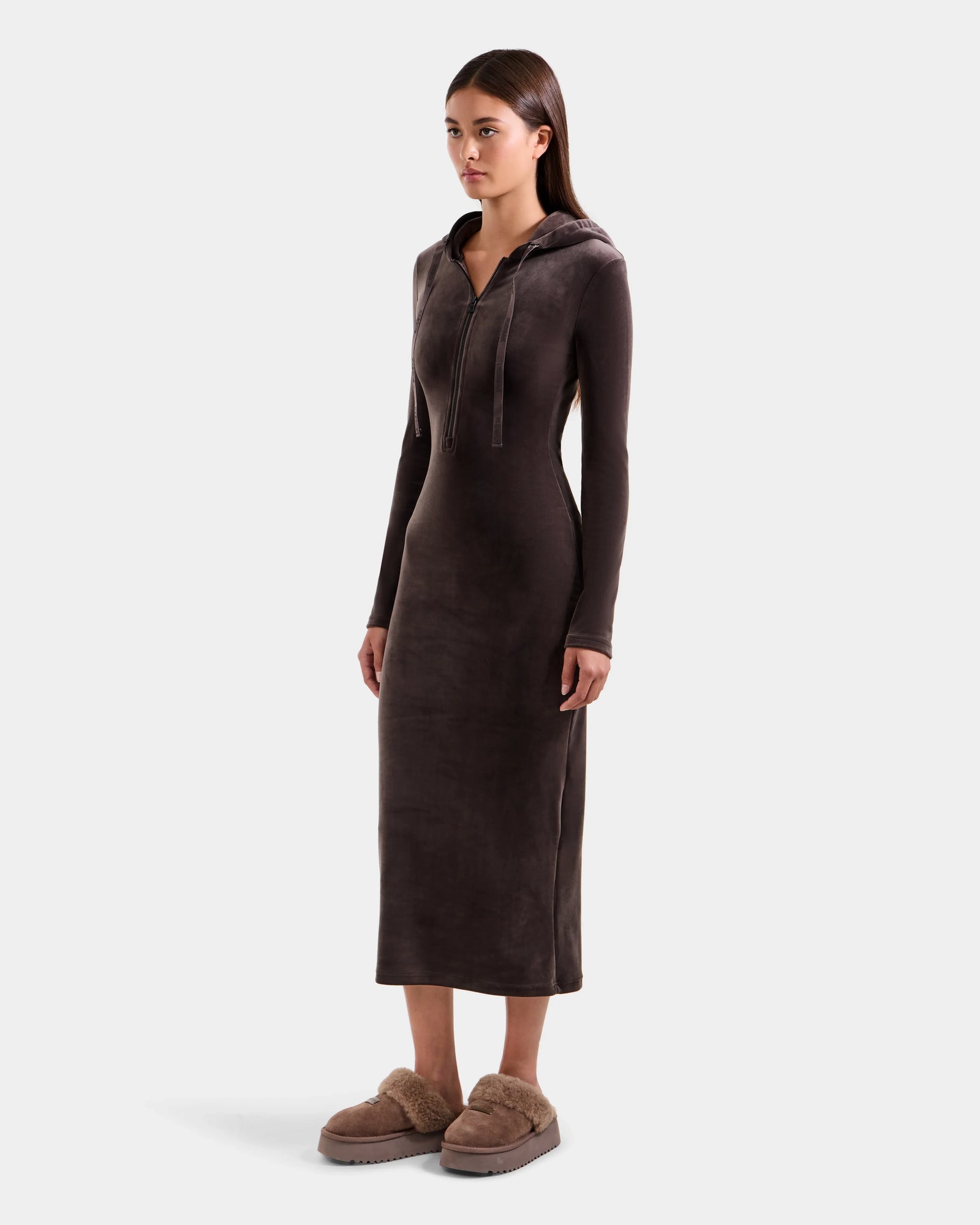 A woman wears the Rudsak ISABELLA - L525502 velour hooded midi dress in Espresso, paired with light brown fuzzy slippers. She stands facing slightly right against a plain light background.