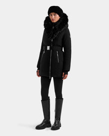 A person stands against a plain light background wearing the Rudsak ISELLE - L125547 Black women’s down parka with a fur-trimmed hood, paired with black leggings, tall black boots, and a black hat.