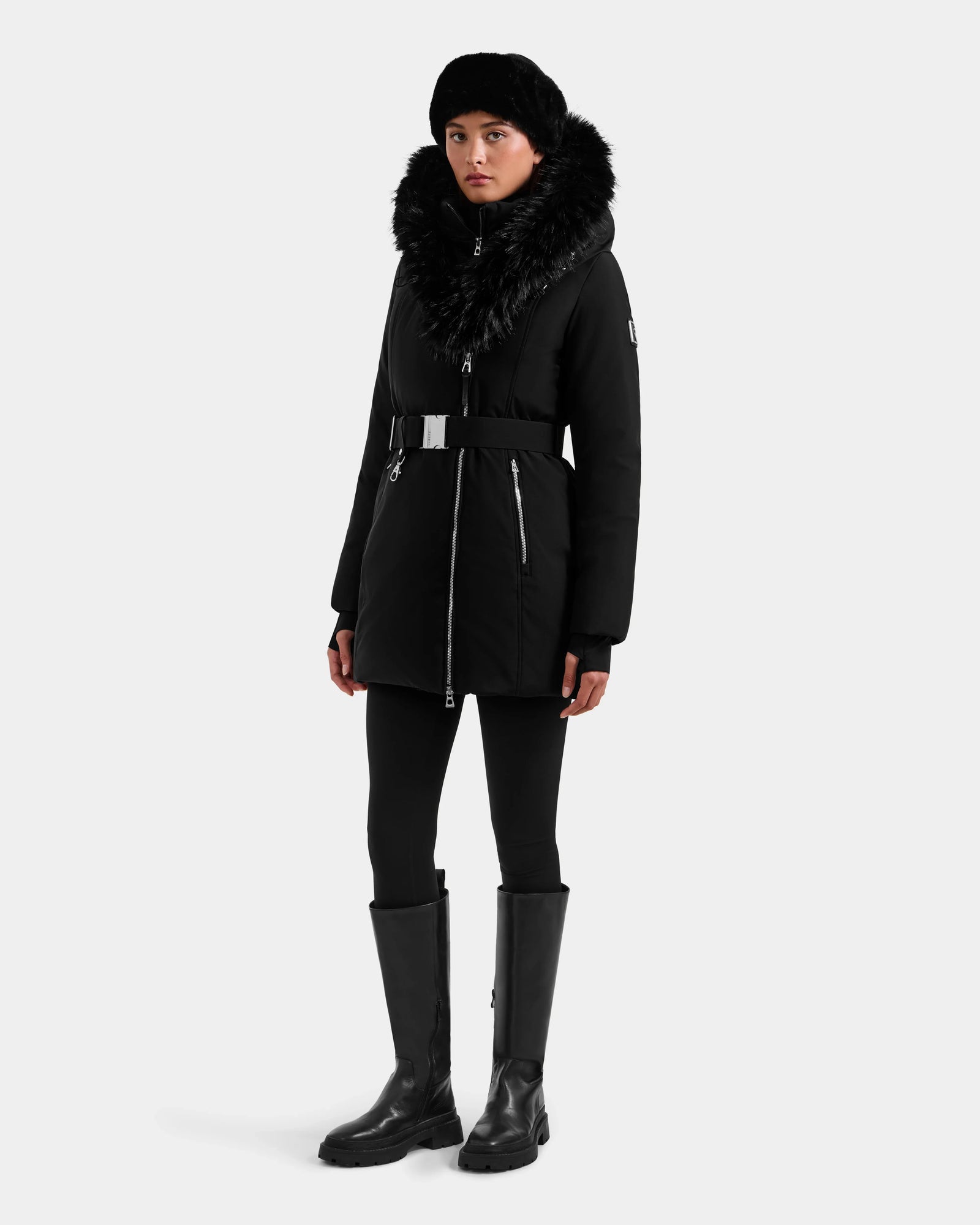 A person stands against a plain light background wearing the Rudsak ISELLE - L125547 Black women’s down parka with a fur-trimmed hood, paired with black leggings, tall black boots, and a black hat.