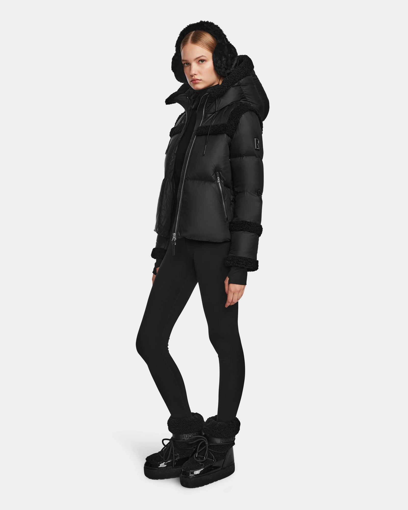 A person wearing the Rudsak JULIANA - L124509 Black/Black women’s short down puffer with RDS-certified down fill, fitted leggings, and winter boots stands against a plain white background.