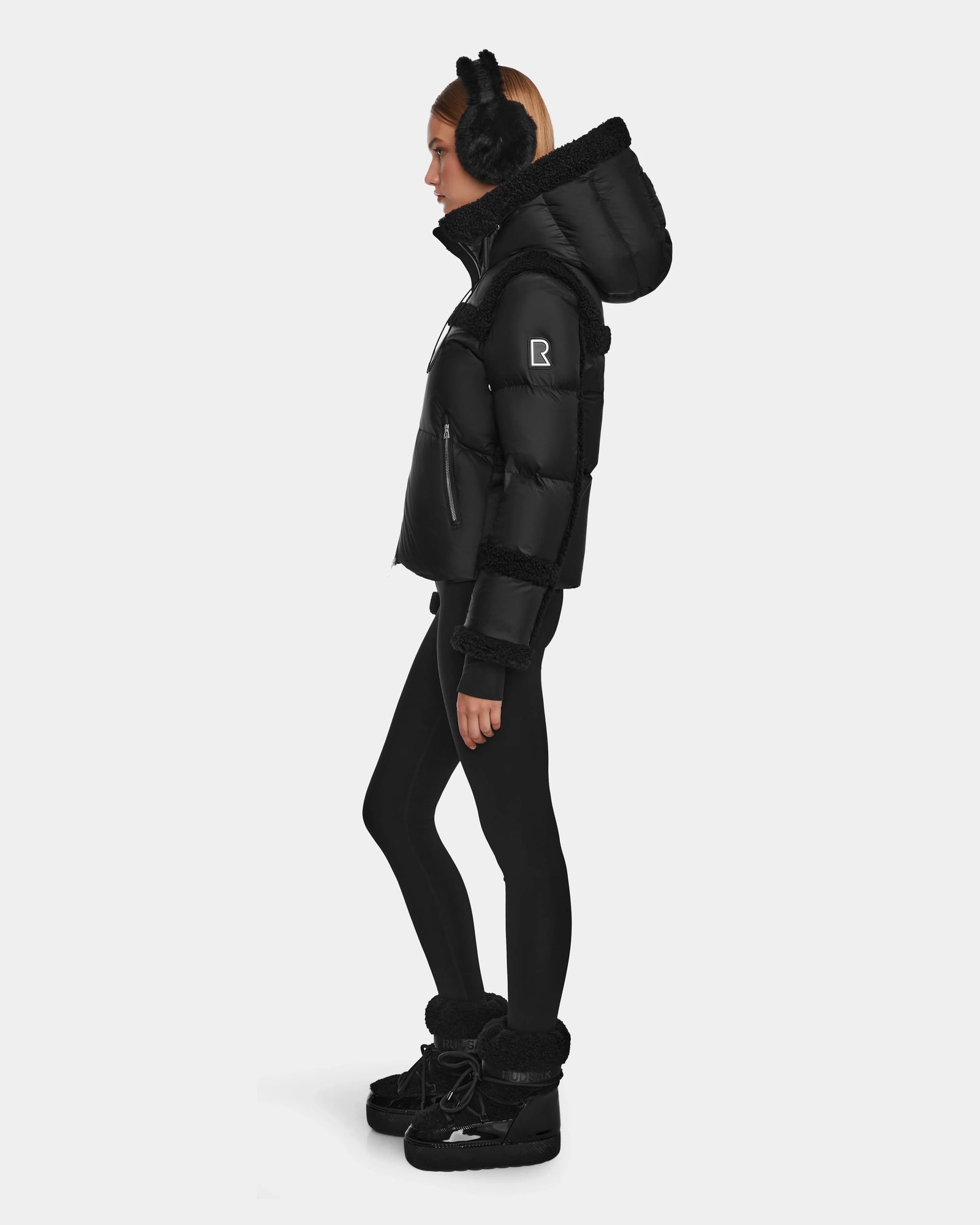 A woman in profile wears the Rudsak JULIANA - L124509 Black/Black women’s short down puffer with RDS-certified down fill, paired with black earmuffs, leggings, and winter boots, standing against a plain white background.