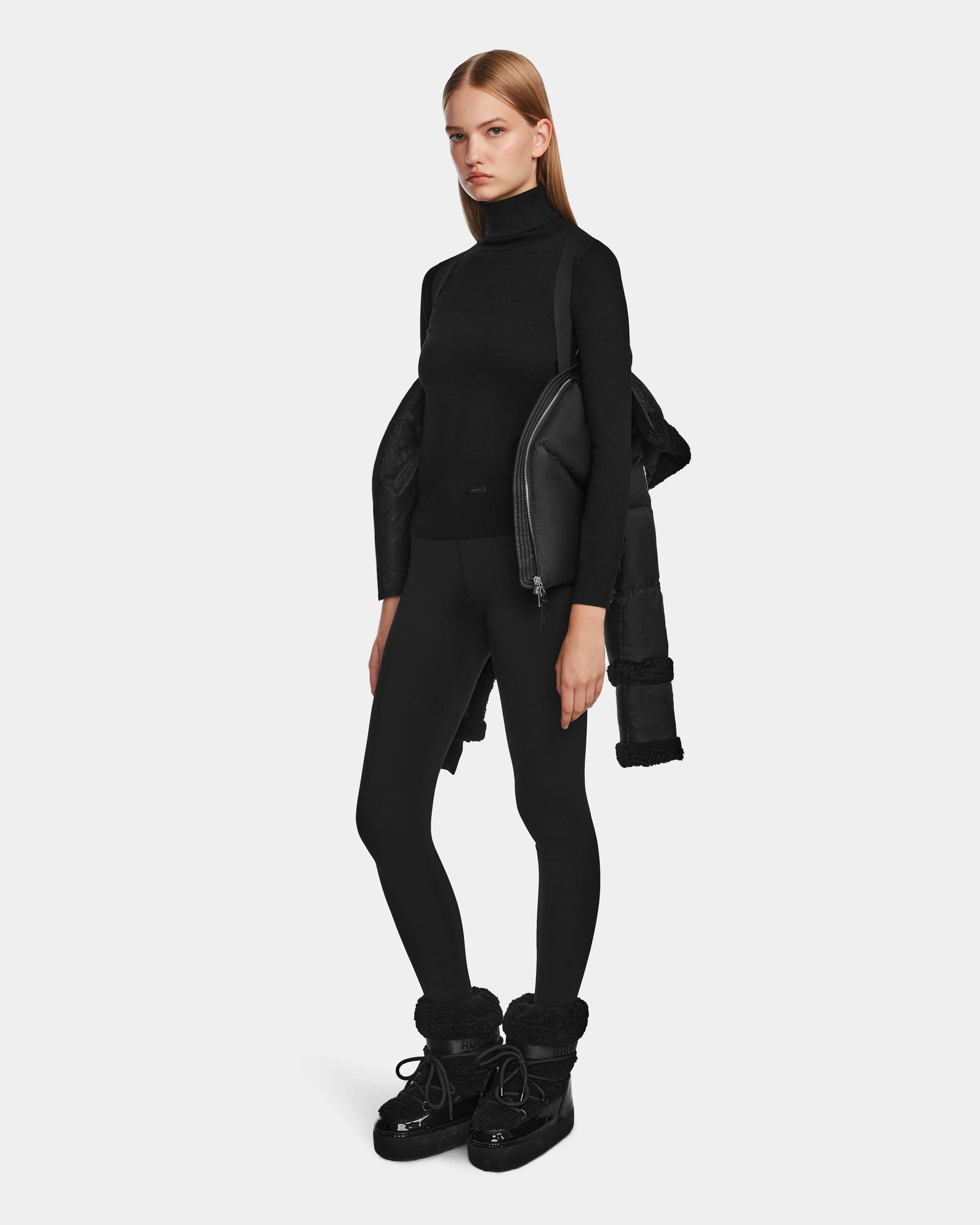 A woman with straight blonde hair in a black turtleneck and leggings holds the Rudsak JULIANA - L124509 Black/Black women’s short down puffer over her shoulder, standing against a plain white background.