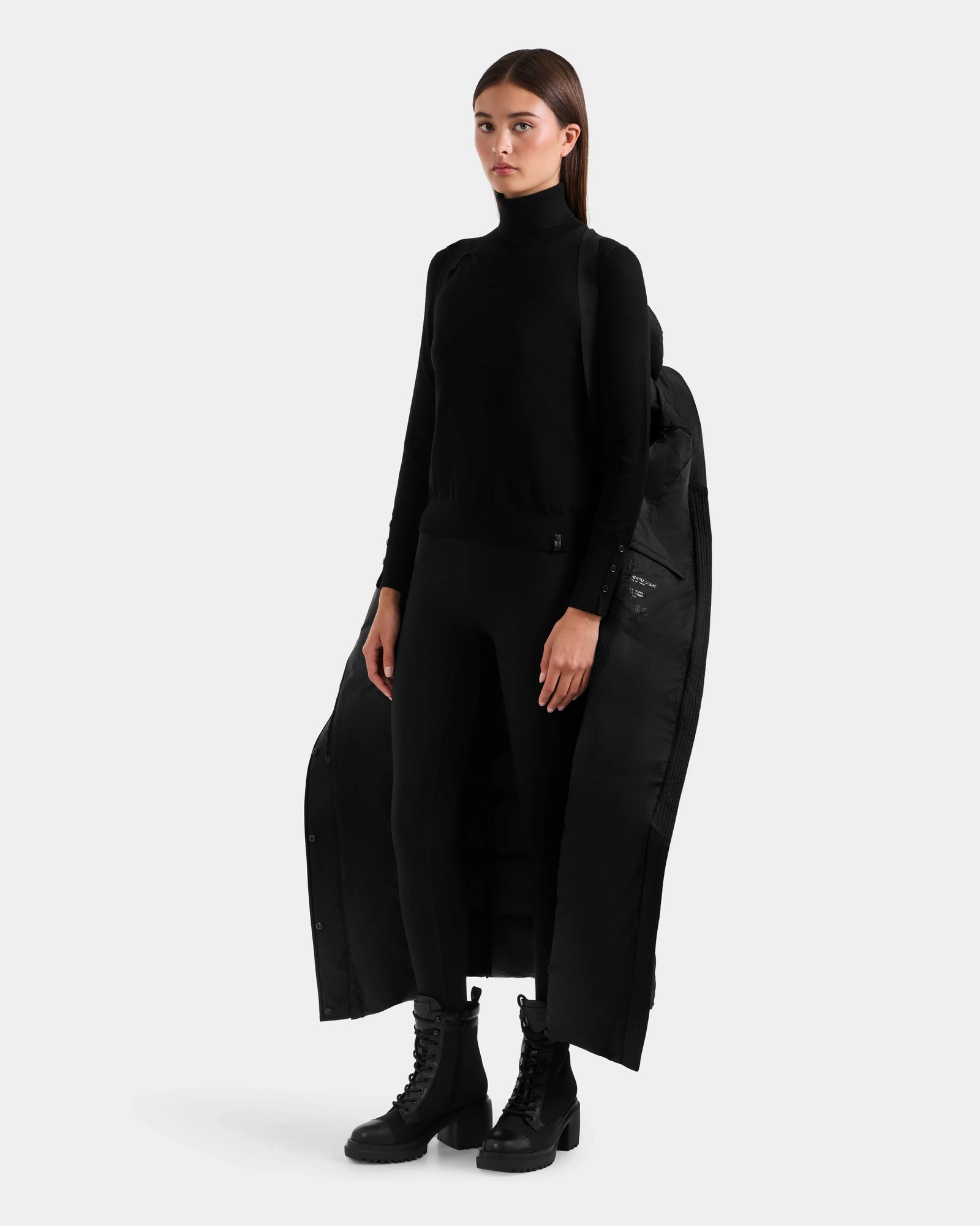 A woman stands against a plain background wearing the Rudsak JULIETA M-L - L125559 Black weather-resistant shell coat draped over her shoulders, paired with a black turtleneck, black pants, and black lace-up boots.