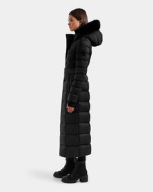 A woman stands in profile against a light grey background, wearing the Rudsak JULIETA M-L - L125559 Black maxi down puffer with a belted waist, black fur-trimmed hood, and black lace-up boots.