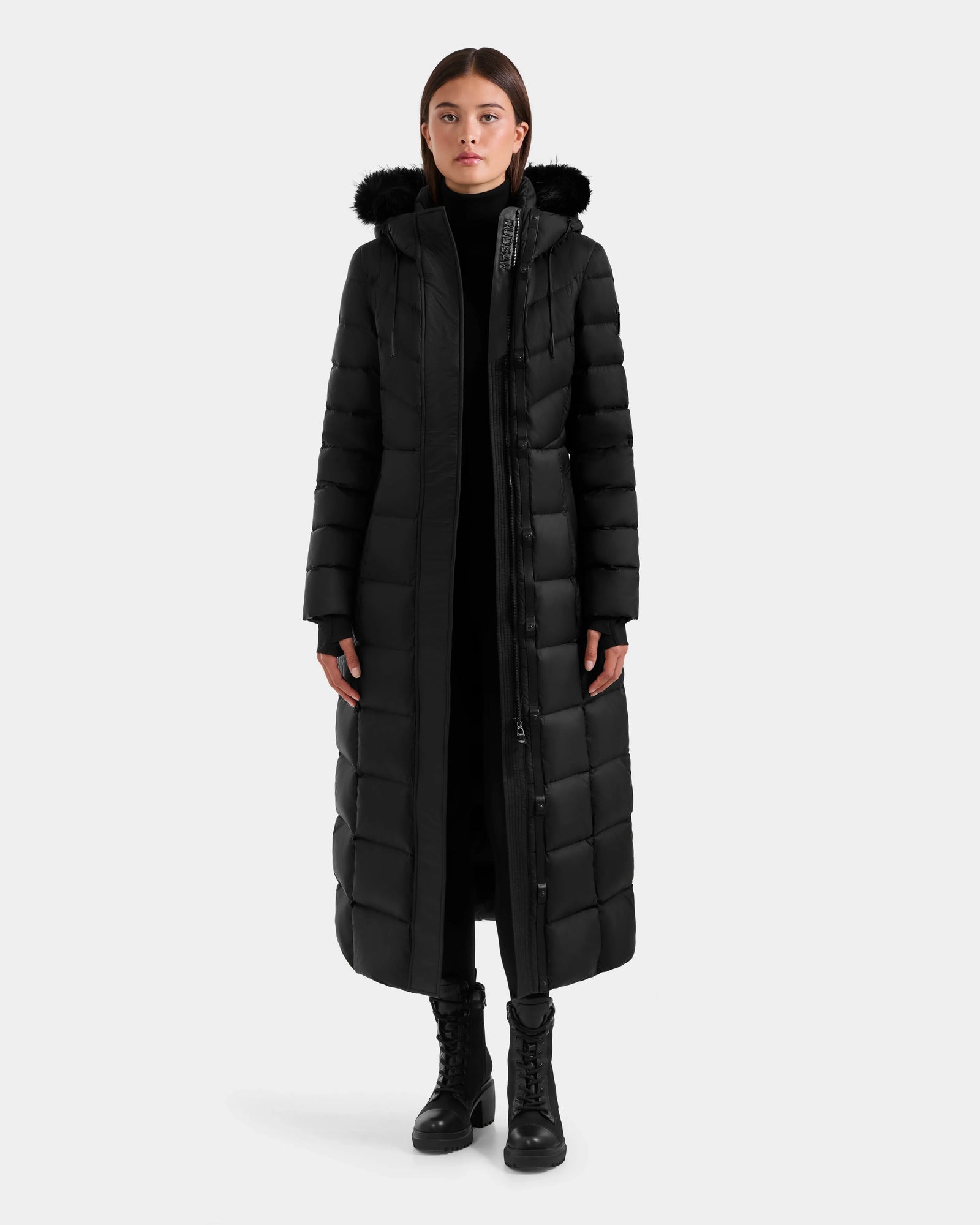A woman faces forward wearing the Rudsak JULIETA M-L - L125559 Black maxi down puffer jacket with a weather-resistant shell and black fur-lined hood, paired with black pants and lace-up boots against a plain light background.